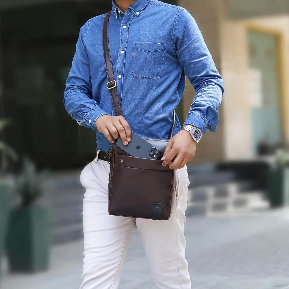 Slim brown leather messenger bag for men with front pocket and long strap, worn crossbody style.