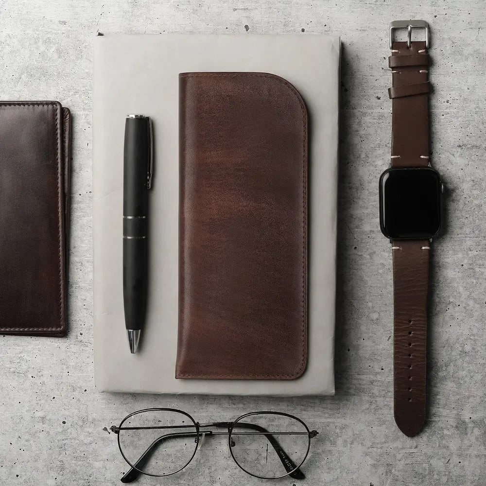 Brown leather glasses pouch placed with watch, pen, and notebook on a desk