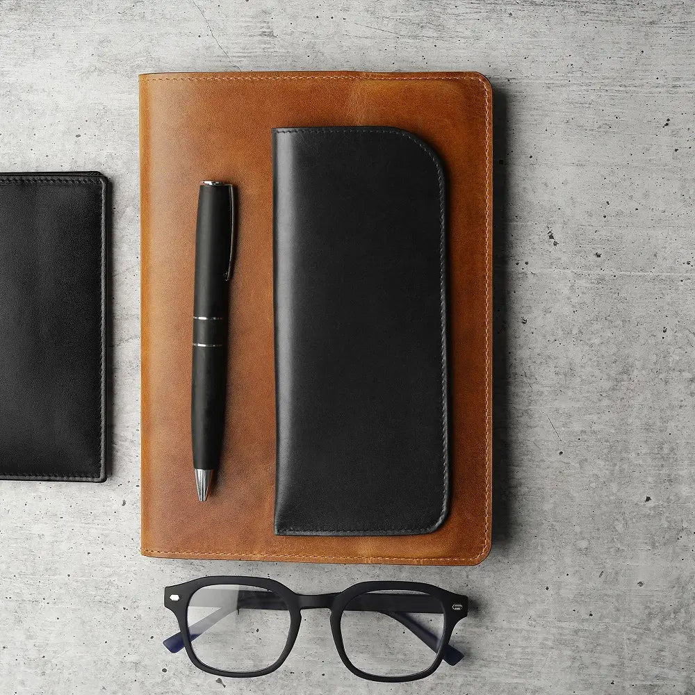 Black leather glasses pouch placed on brown leather notebook with pen and spectacles