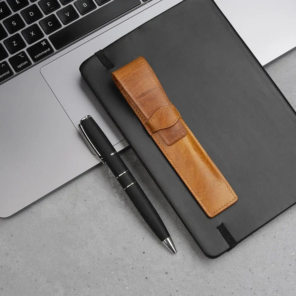 Tan leather pen holder placed neatly on an open diary beside a pen and laptop, styled for a modern desk setup.