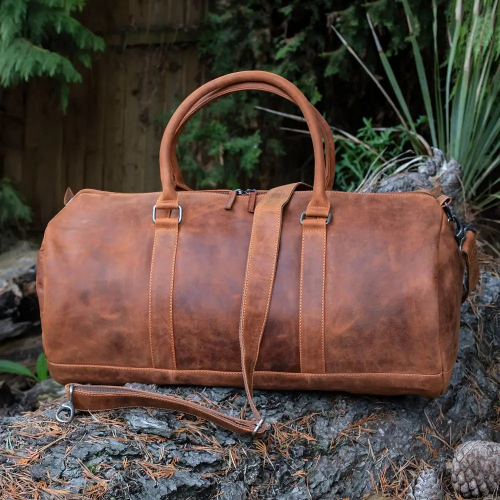 Vintage tan leather holdall placed outdoors on a tree trunk, showing its sturdy handles, detachable shoulder strap, and classic duffel design.