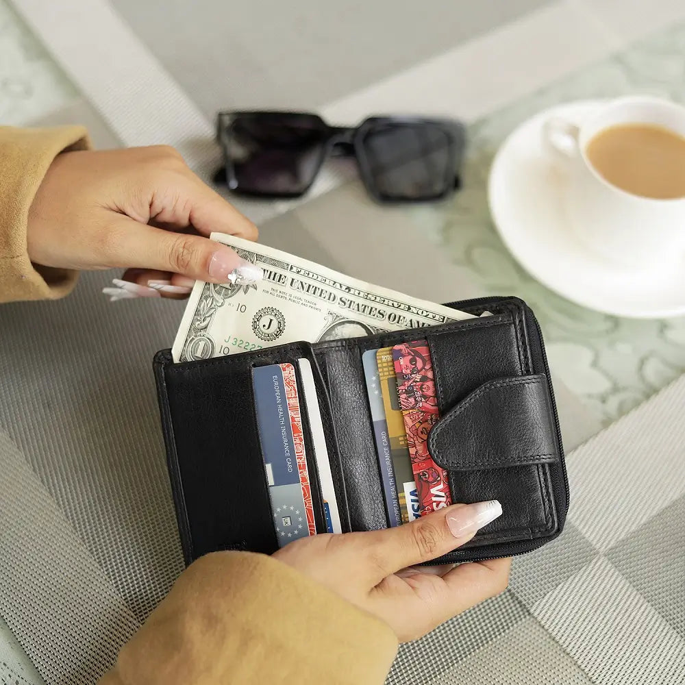 Person holding a black wallet with money and cards, with a cup of coffee and sunglasses in the background.