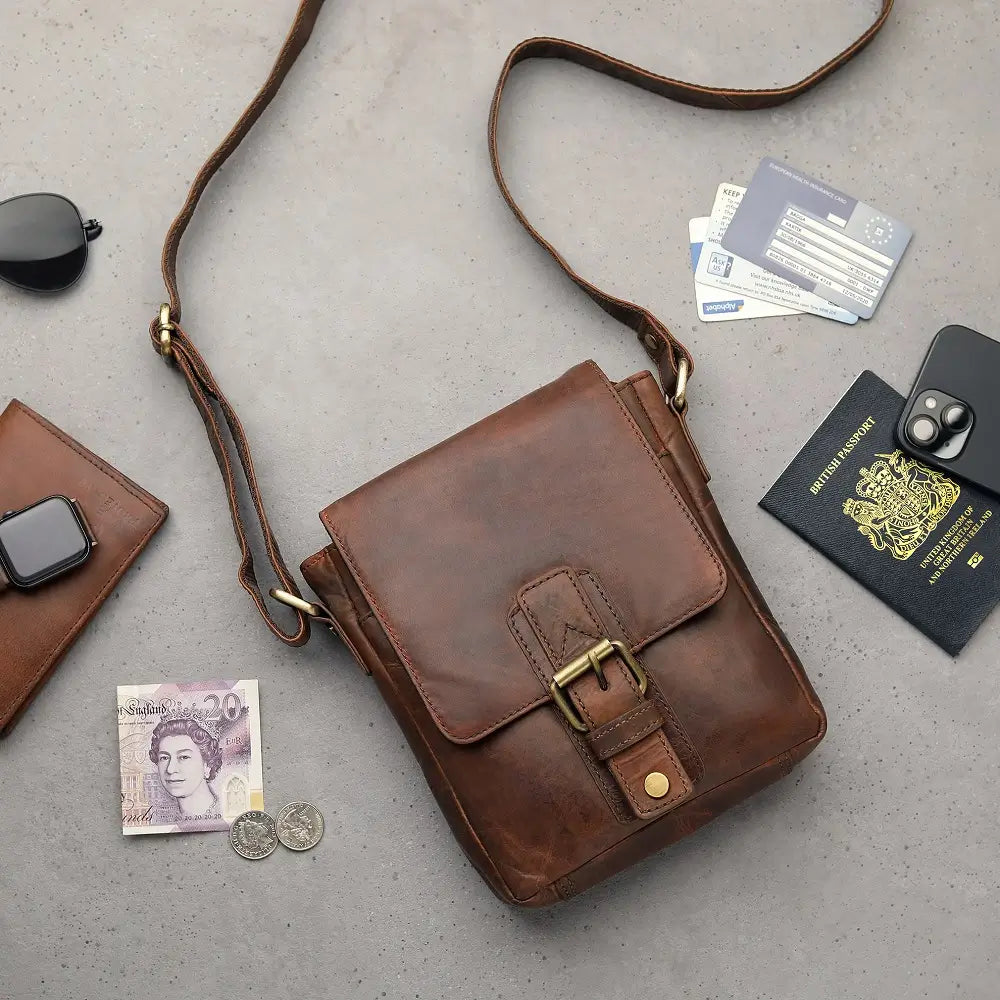 Brown leather bag with accessories including a passport, phone, and money on a gray surface