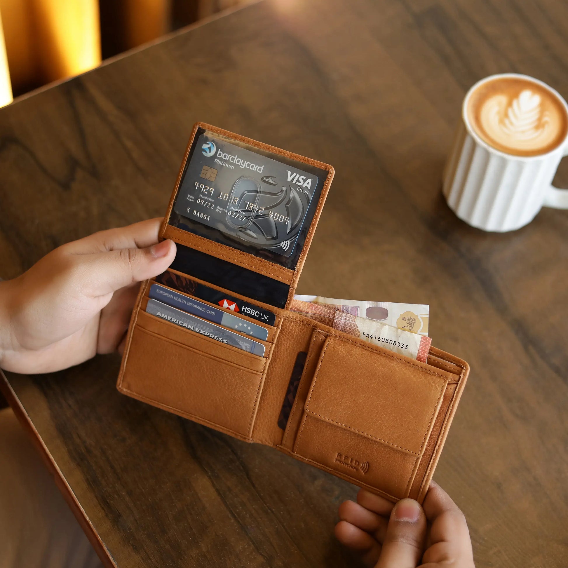 Cognac leather wallet with cards and cash held by a hand on a wooden table with a coffee cup in the background.