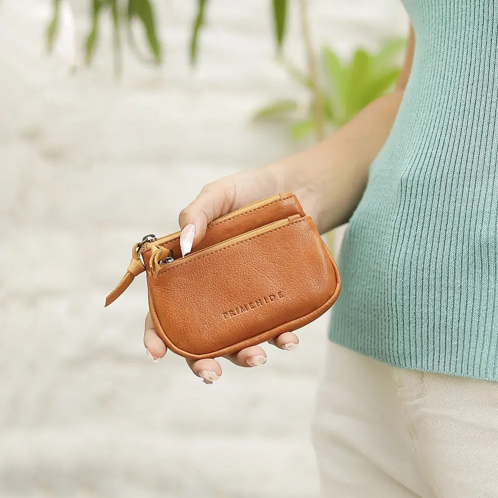 Person holding a Cognac leather pouch with 'Trimenhide' branding against a blurred natural background