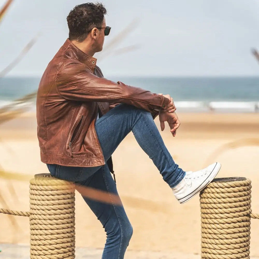 Man in cognac leather jacket and jeans sitting on a rope barrier by the sea.