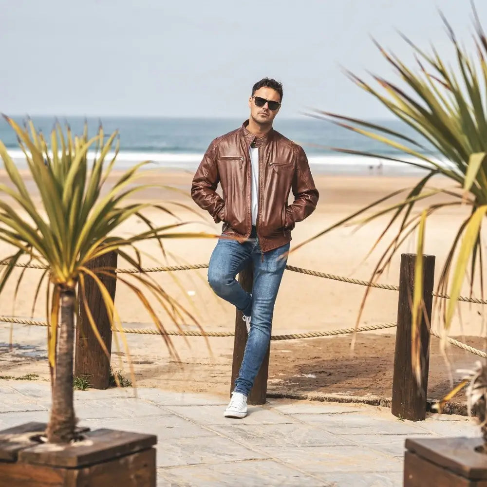 Man in a cognac leather jacket and jeans standing on a beach with palm trees.