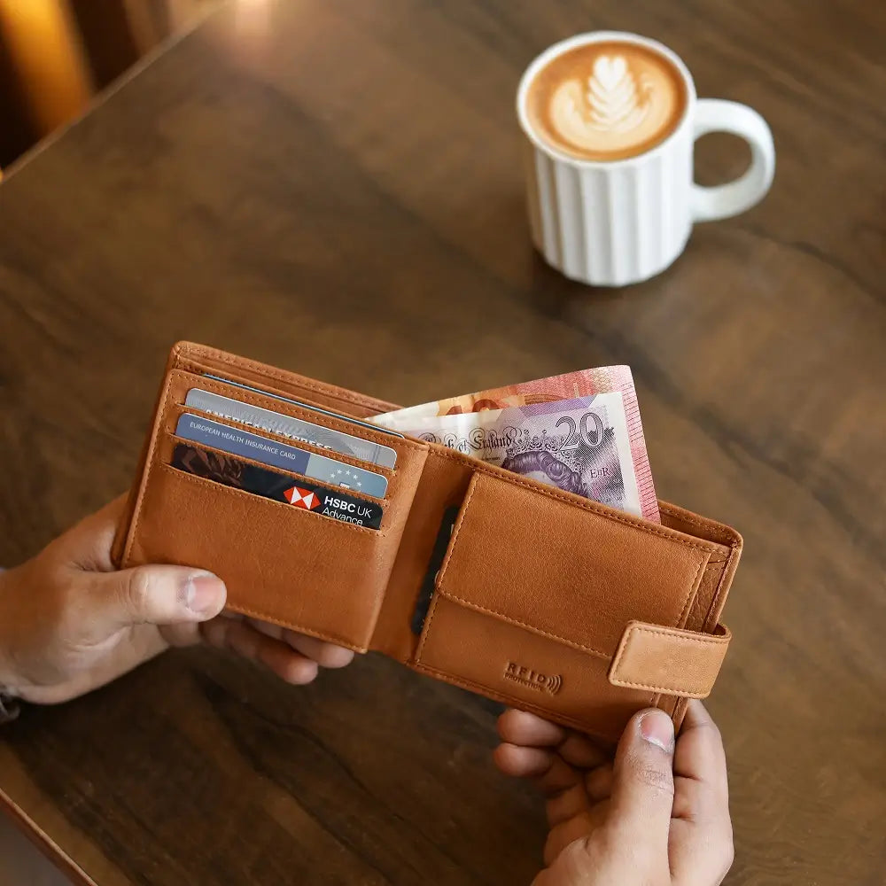 Cognac leather wallet with money and cards held by hands on a wooden table with a coffee cup in the background.