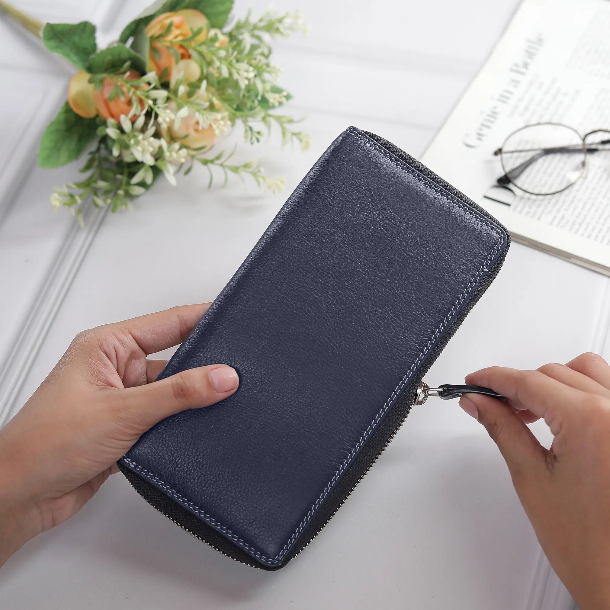 Person opening a Navy purse with a white surface, glasses, and flowers in the background