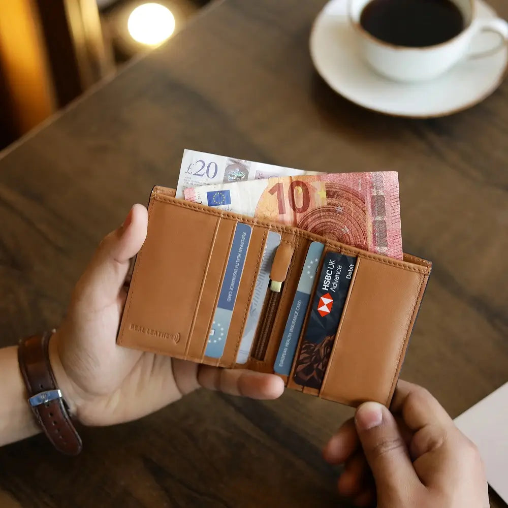 Tan leather wallet with money and cards held by a person, with a cup of coffee in the background.