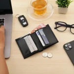 Flat lay of Basic Wallet Style 87 black leather RFID bifold wallet with tab closure, shown with coins, laptop, and glasses