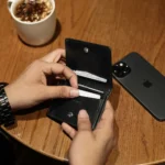 Black leather Mayfair 2511 credit cardholder being used at a café table beside a smartphone and coffee.