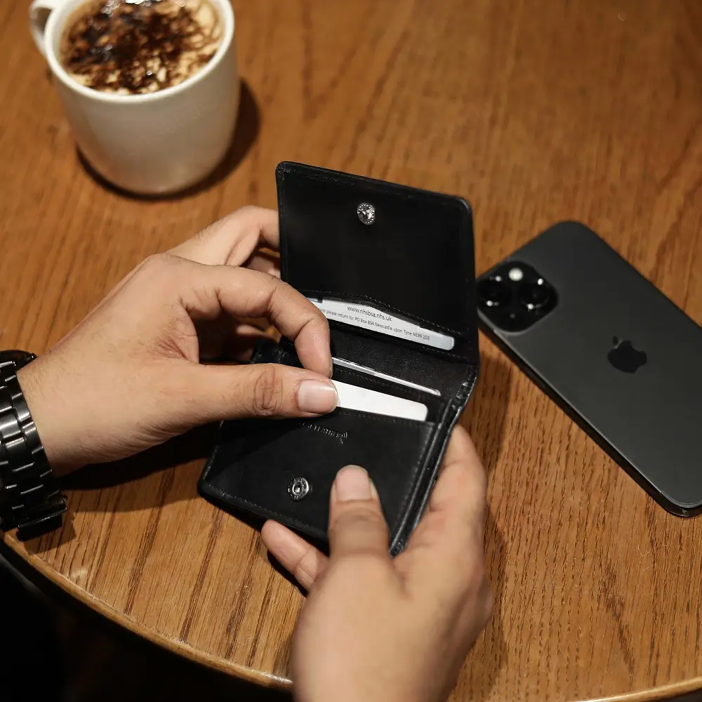 Black leather Mayfair 2511 credit cardholder being used at a café table beside a smartphone and coffee.