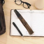 Brown leather pen holder placed on an open diary with a pen and glasses, styled for a modern workspace.